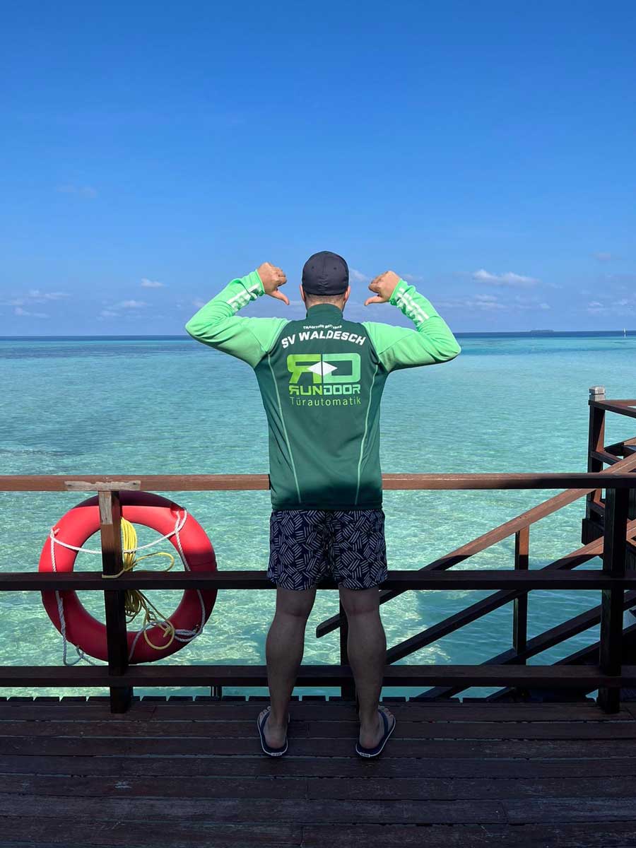 Trikot auf Tour - Mann am Meer mit Blick von einer hölzernen Balustrade auf auf das türkisblaue, flache Wasser